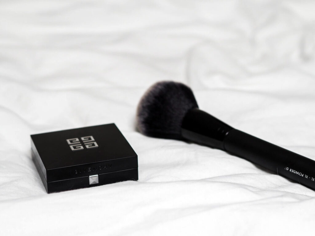 small makeup compact on a bed with a fluffy makeup brush next to it